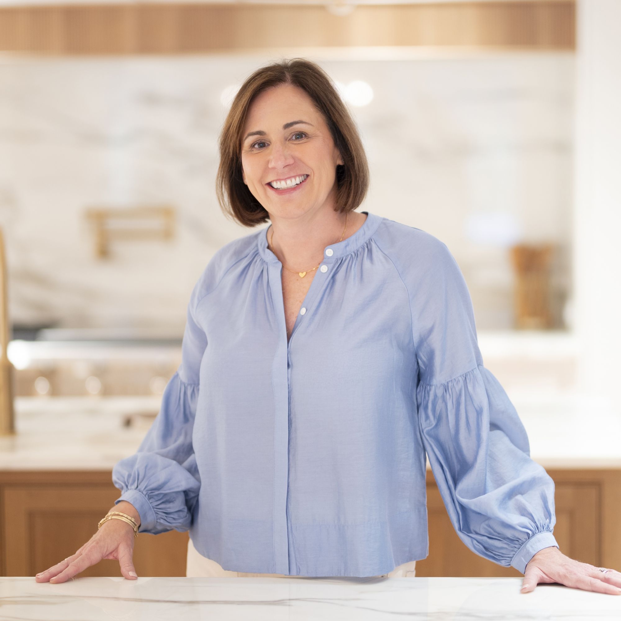 A headshot of Ashley Bailey wearing a blue shirt and white trousers against a blurred kitchen background
