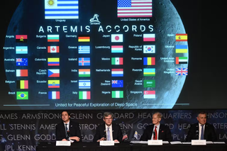 Four men discussing Artemis Accords in front of a screen showing flags in front of the moon.