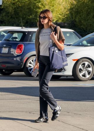 Kaia Gerber wearing a graphic tee with a blue tote bag, pants, and Adidas Sambas