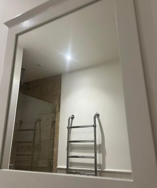 A mirror with a white wooden border, and silver towel rail, glass shower door, brown tiles and overhead shower in the reflection