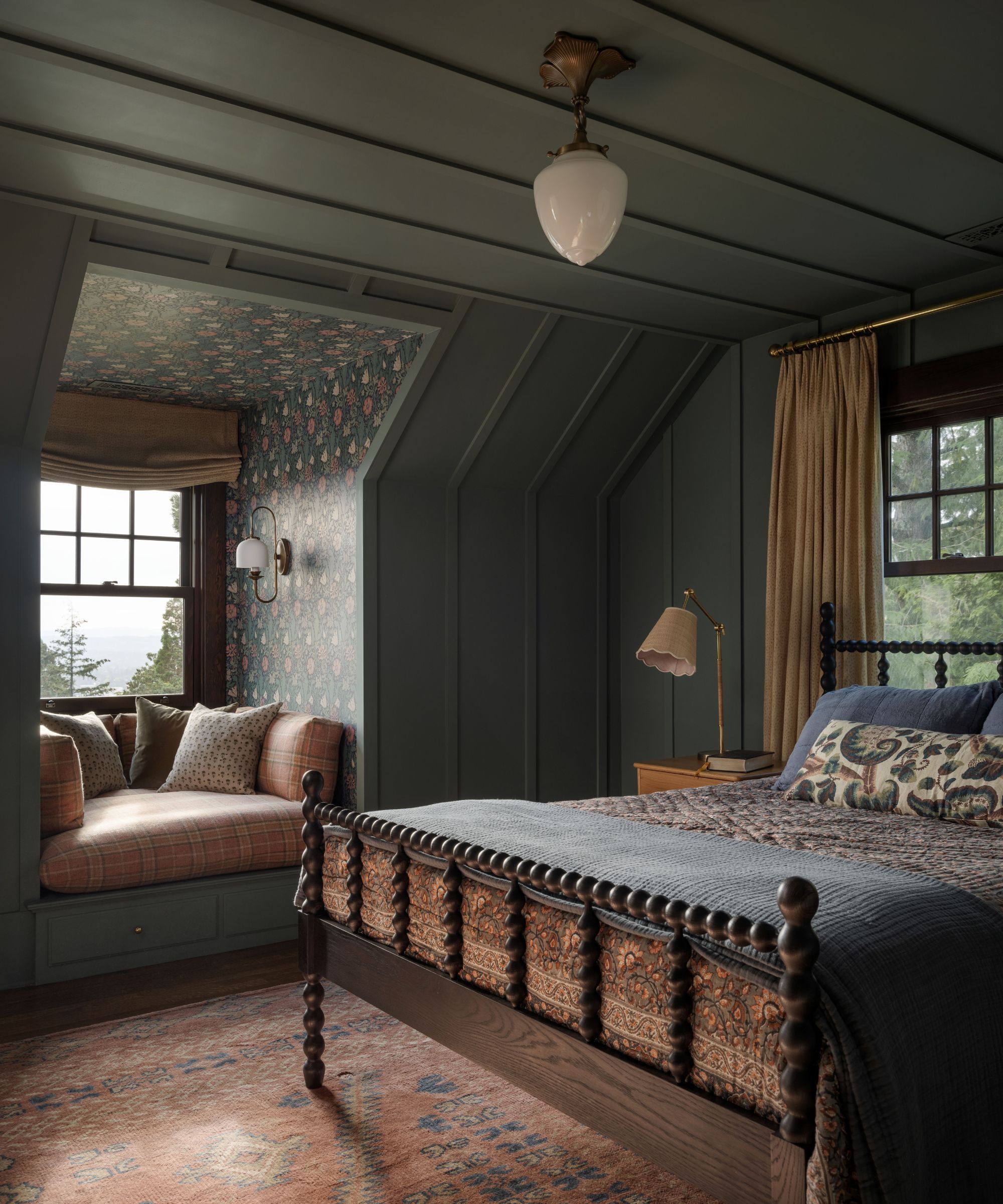 Autumnal bedroom with dark grey walls, a bobbin wood bed with dark blue bedding, a pink Persian rug on the floor, and a built-in window seat with pink plaid cushions in a wallpapered alcove.
