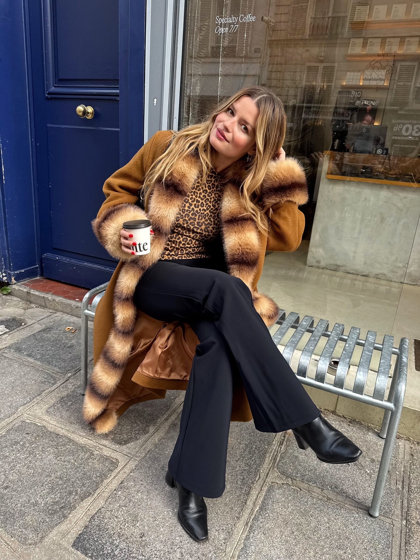 An image of an influencer in Paris sitting on a bench wearing a brown coat with striped faux fur-trim, black wide-leg trousers and black leather boots.