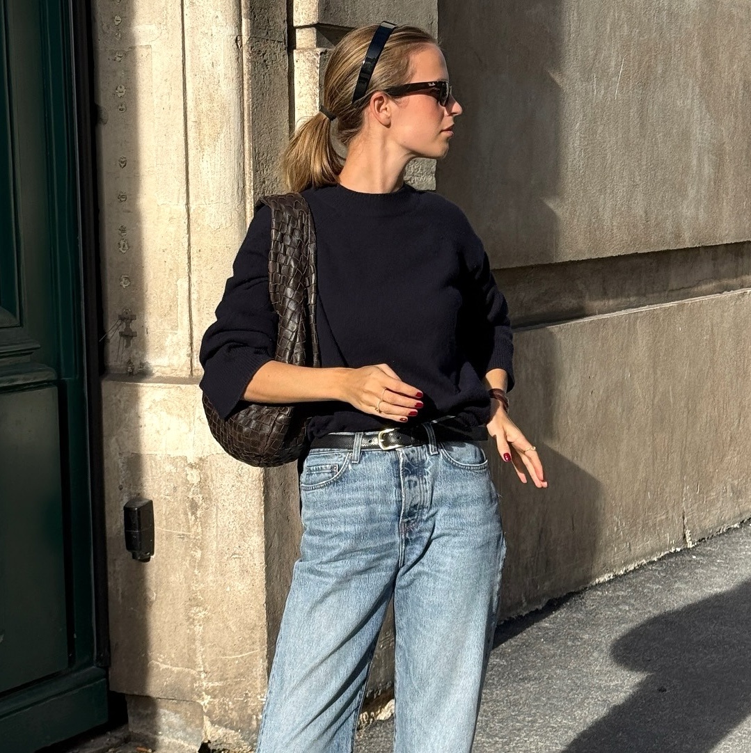 Danish fashion creative Clara Dyrhauge poses on a Paris sidewalk wearing a black headband, sunglasses, a black crewneck sweater, woven brown Bottega Veneta shoulder bag, a thin black belt, and jeans