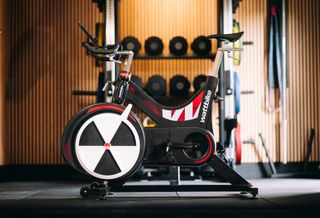 Wattbike Air with its black and wheel air restiance cover and trackmark red logo. Sat in a gym location featuring dunbells and weights