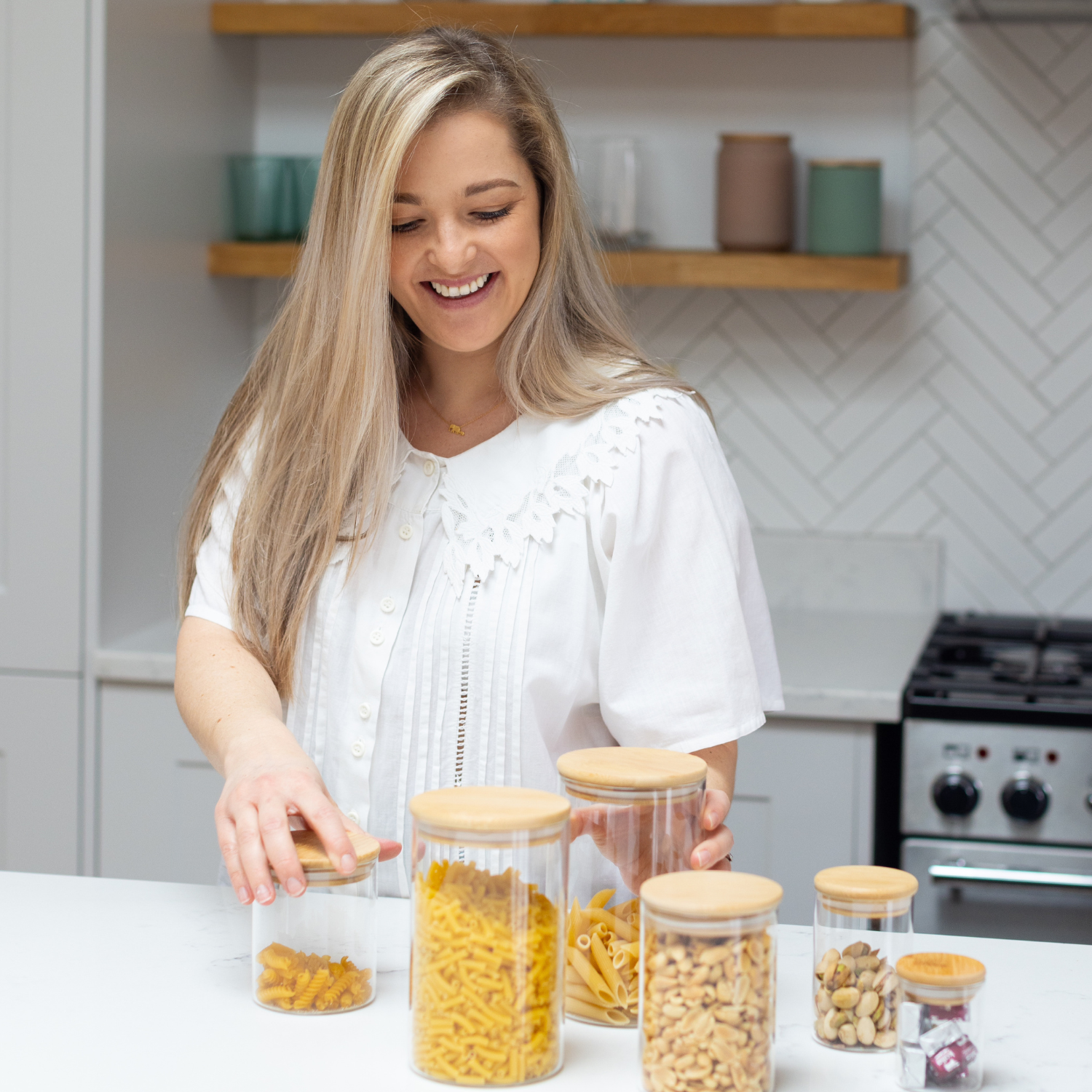 A woman with straight blonde hair in a white top organizing pasta into glass jars with wood lids