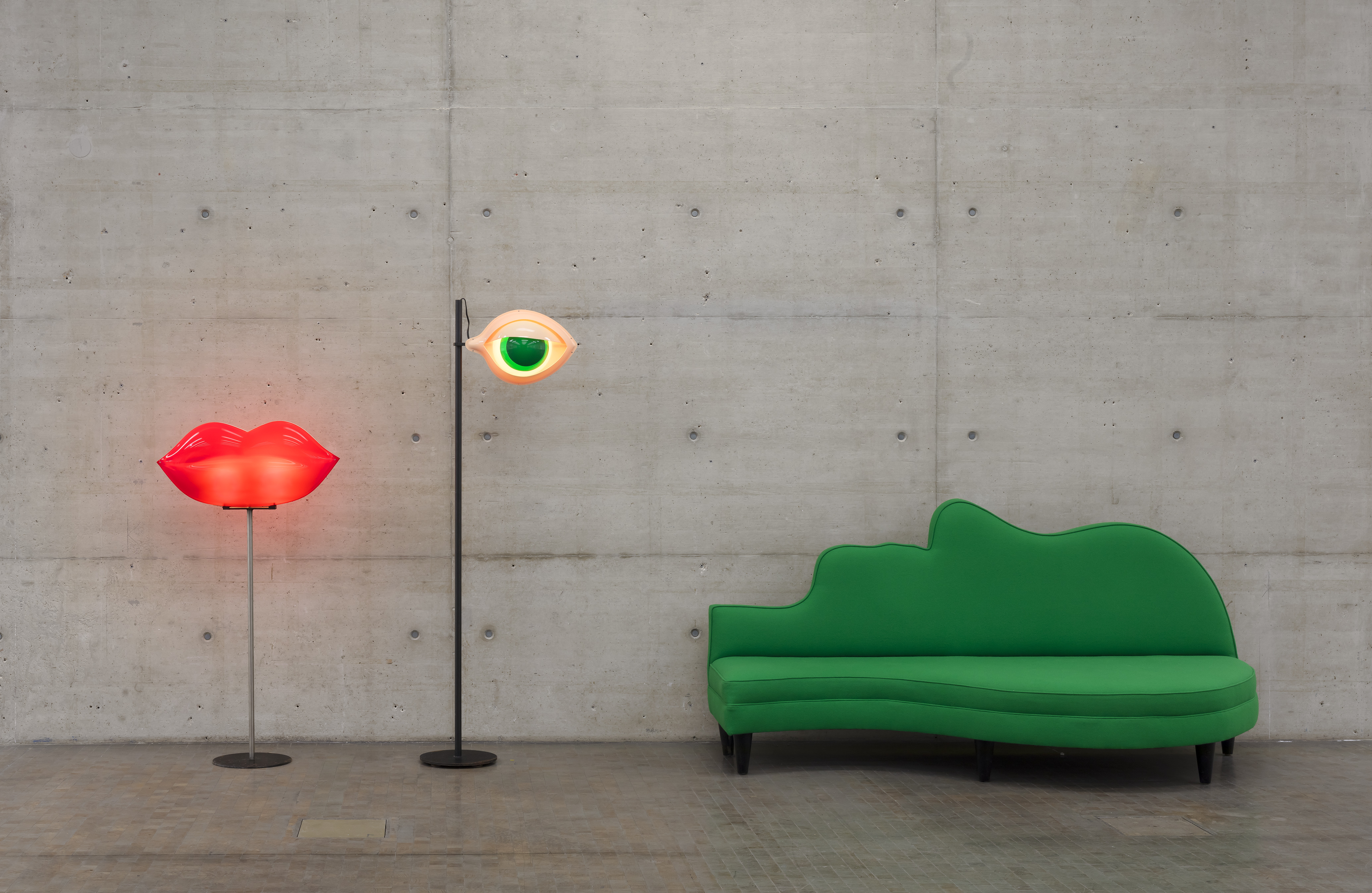 A standing lamp shaped like bright red lips, on a chrome pedestal, one shaped like a green eye, and a sofa shaped like a human face seen sideways in emerald green sit in a brutalist room with concrete walls and floors.