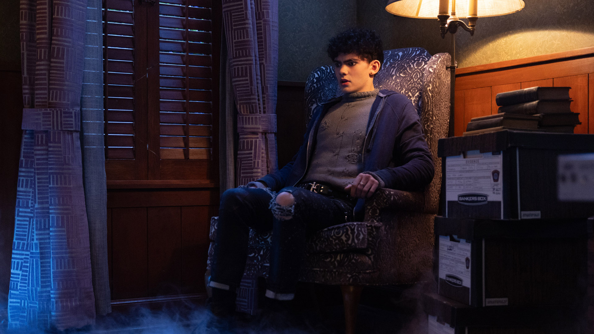 Teenager sits on a chair at night with a lamp next to him in Agatha All Along