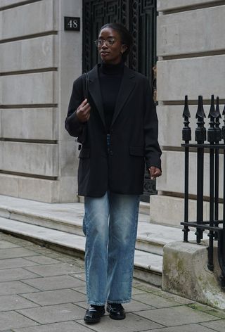 A black turtleneck outfit is shown in a photo of a woman standing on the sidewalk wearing a black blazer over a black turtleneck with relaxed jeans and black loafers