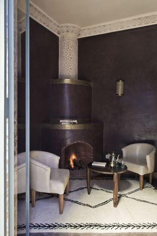 A hotel sitting area with white chairs, a round wooden coffee table, a fireplace, a white black lined rug, a wall light and dark grey walls.