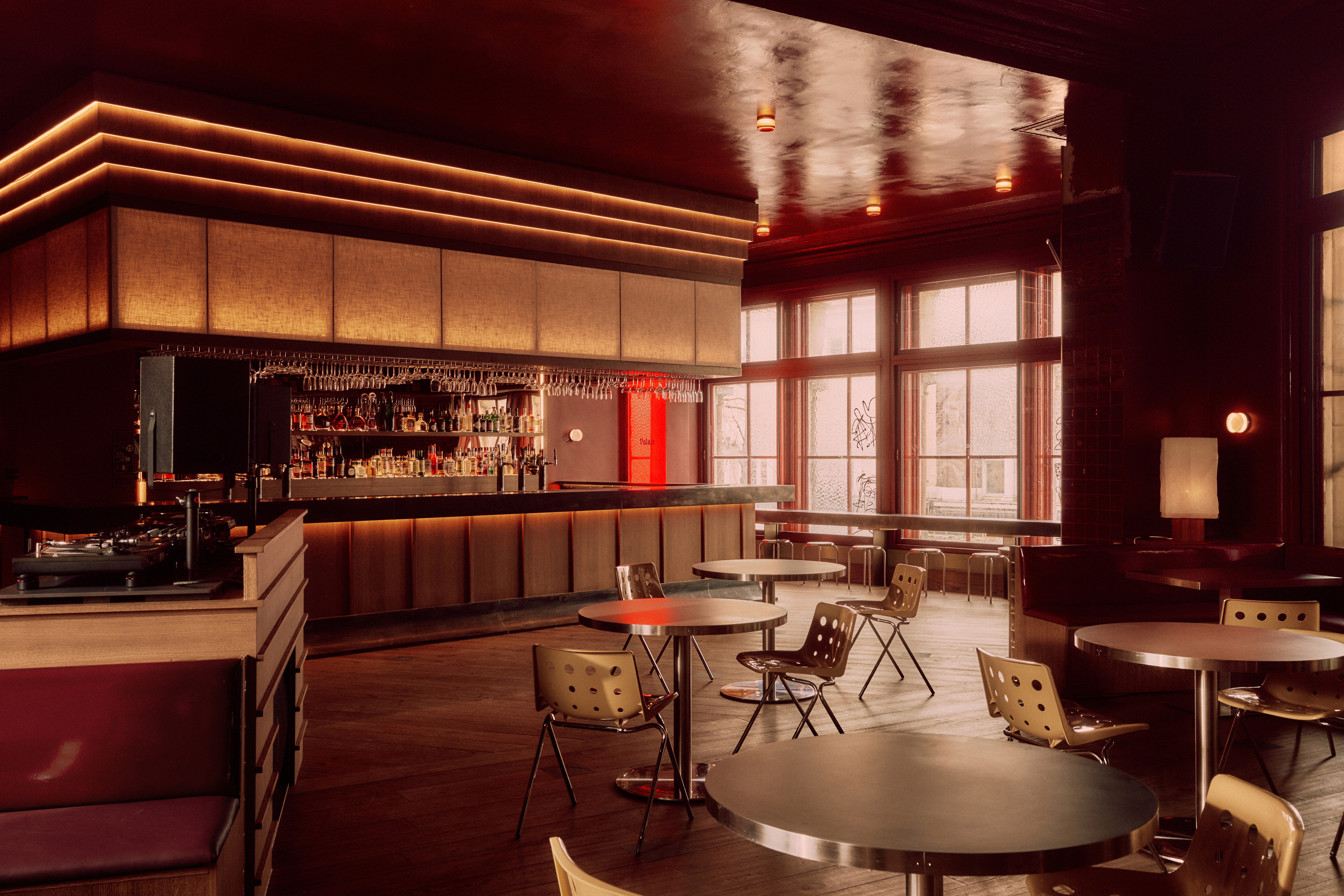 A red-tinted bar with a sleek wooden countertop, back-lit surfaces, plenty of drinks stocked in its back, and a red, rectangular sign reading "Palais".