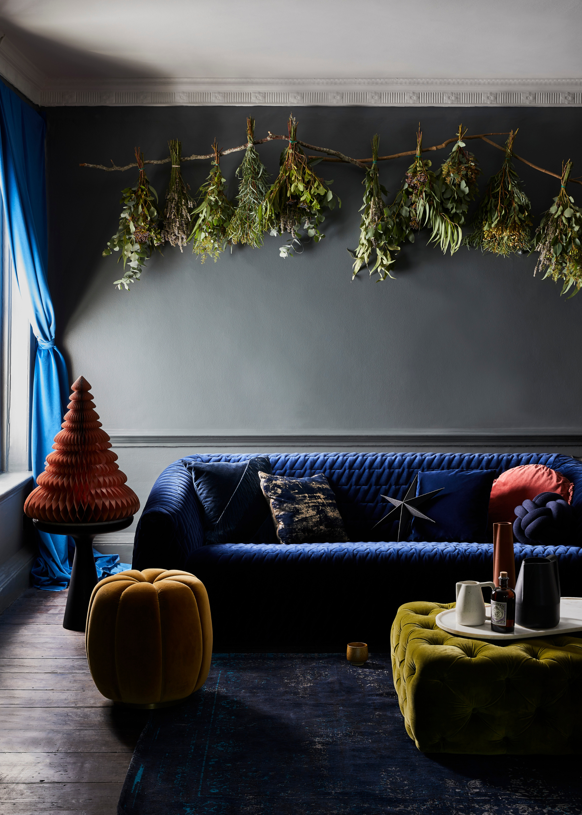 A dark gray-hued wall with blue curtains and a blue sofa has a unique branch display with upside-down hanging tied foliage that look like mini Christmas trees displayed at the top of the wall with a red paper decoration on a side table