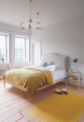 white bedroom with yellow rug and bedspread, upholstered bed, white walls, wooden floorboards and decorative ceiling light