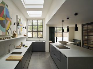 a side returned extension with a grey kitchen. In the kitchen is a large island and worktops. Above the worktop is open shelving with hanging artwork.