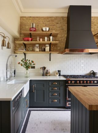 Dark green kitchen cabinetry with white countertops and backsplash, exposed brick and open shelving.