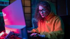 Young female hacker wearing a hooded sweatshirt working on a desktop computer in a dimly lit room with glowing lights reflecting on back of computer monitor.