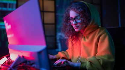 Young female hacker wearing a hooded sweatshirt working on a desktop computer in a dimly lit room with glowing lights reflecting on back of computer monitor.