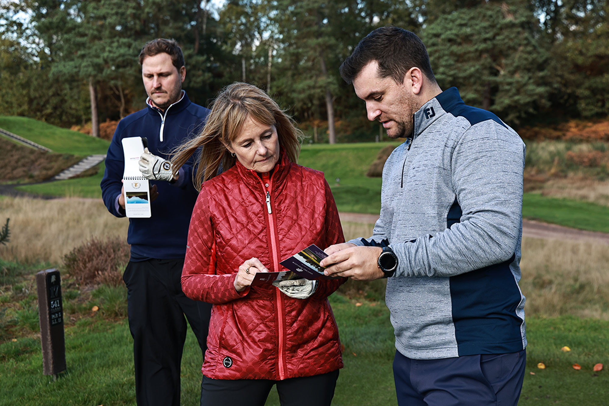 Three players reviewing their scorecards and discussing the outcome, with one player showing his scorecard to another player and explaining a particular element as the other player watches on