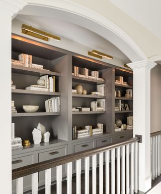 Landing space with large built-in bookcase, painted in a dark brown shade. Shelf is decorated with a collection of ornaments, books and decorative accessories, all in a neutral color palette.