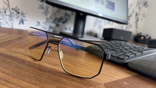 Close-up of Gunnar Novato Onyx computer glasses featuring amber lenses and a matte black geometric metal frame resting on a wooden desk in front of a monitor and keyboard.