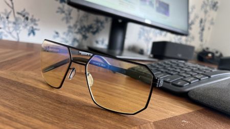 Close-up of Gunnar Novato Onyx computer glasses featuring amber lenses and a matte black geometric metal frame resting on a wooden desk in front of a monitor and keyboard.