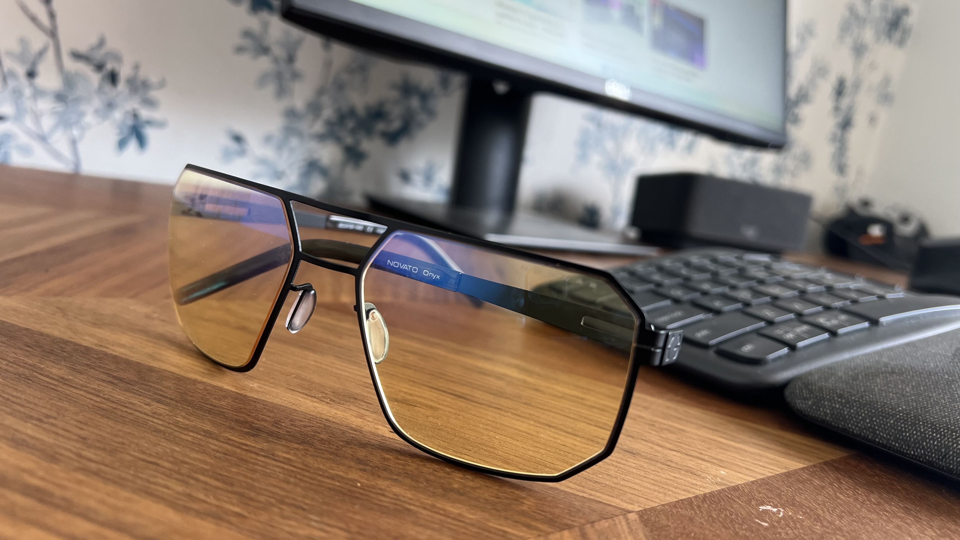Close-up of Gunnar Novato Onyx computer glasses featuring amber lenses and a matte black geometric metal frame resting on a wooden desk in front of a monitor and keyboard.