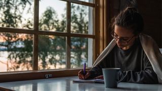 Woman sitting down at desk under blanket with cup of tea journaling