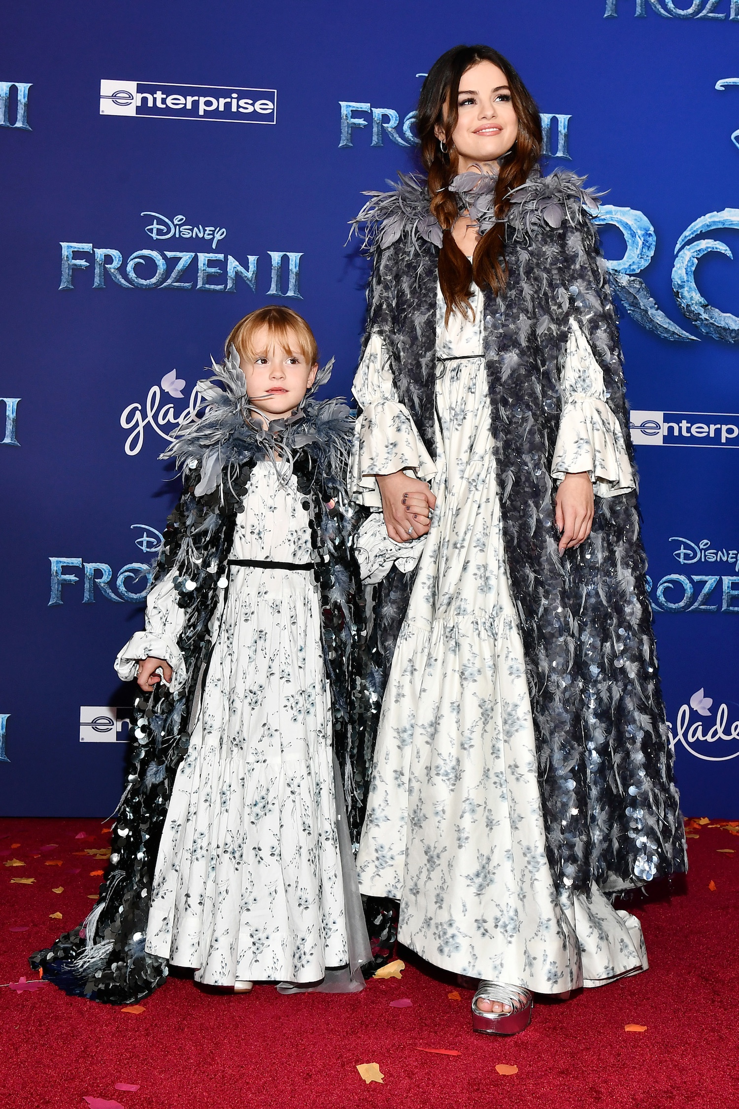 Gracie Elliot Teefey and Selena Gomez wearing matching white printed dresses and capes covered in gray feathers at the premiere of Frozen II in 2019