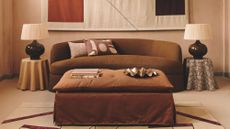A warm, terracotta-toned living room featuring a curved chocolate-brown sofa and a large, abstract geometric wall hanging. A rust-colored ottoman sits in the center on a patterned rug, flanked by two table lamps with pleated shades.