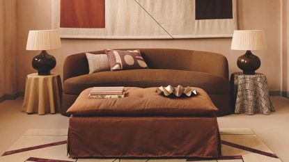 A warm, terracotta-toned living room featuring a curved chocolate-brown sofa and a large, abstract geometric wall hanging. A rust-colored ottoman sits in the center on a patterned rug, flanked by two table lamps with pleated shades.