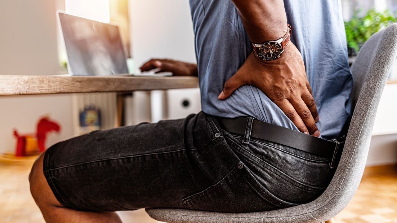 Man supports his lower back with hand as he sits at a table doing work