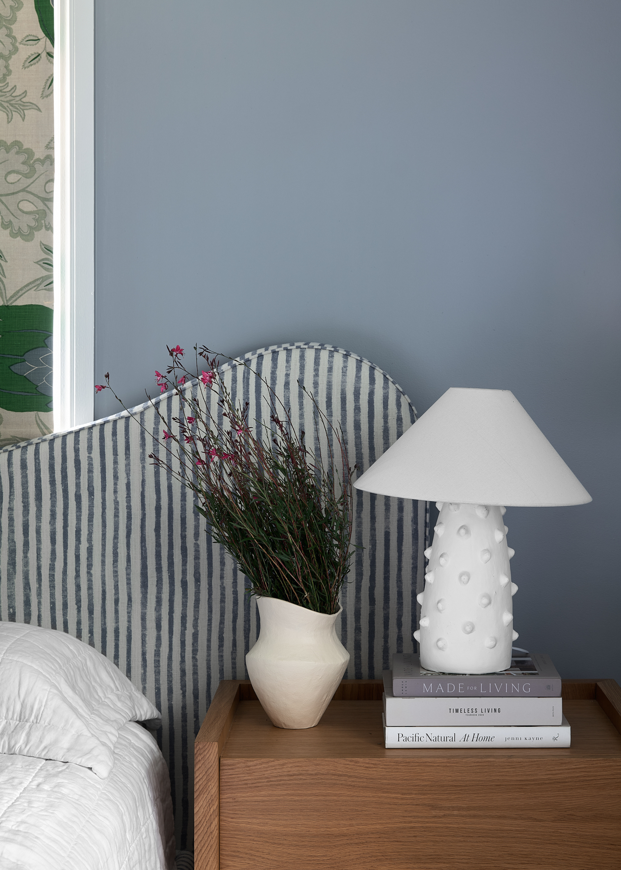 A pale blue bedroom with a striped headboard by a wood nightstand with a white table lamp, a vase of stems, and a stack of books