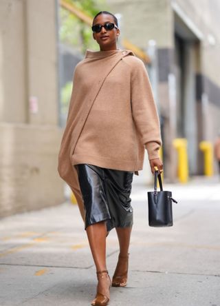 A woman wearing black sunglasses, a light brown draped sweater, shiny black leather jorts, shiny black leather bucket bag and shiny dark brown Dries Van Noten leather heels.