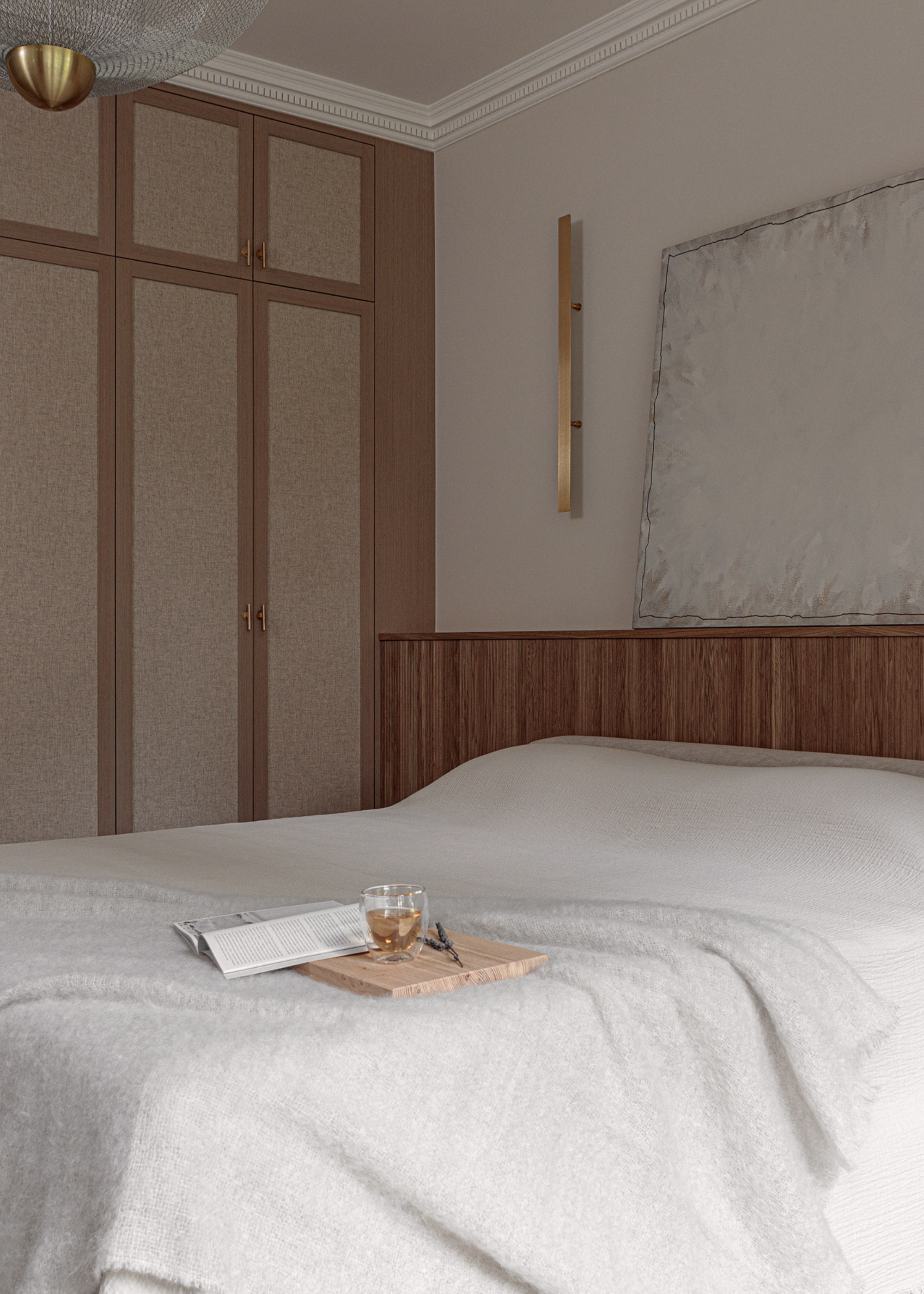 A white bedroom with gold wall sconces, a leaning painting, a bed with a white duvet, a textured white throw and a wooden serving tray with a glass of tea and a book