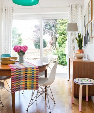 a dining room with a round dining room table with a colorful table runner and pink flowers on it, a white chair, a wall with a glass window door looking onto a garden, a white lamp, a wooden storage unit with a plant and fruit bowl on, and a colorful stool