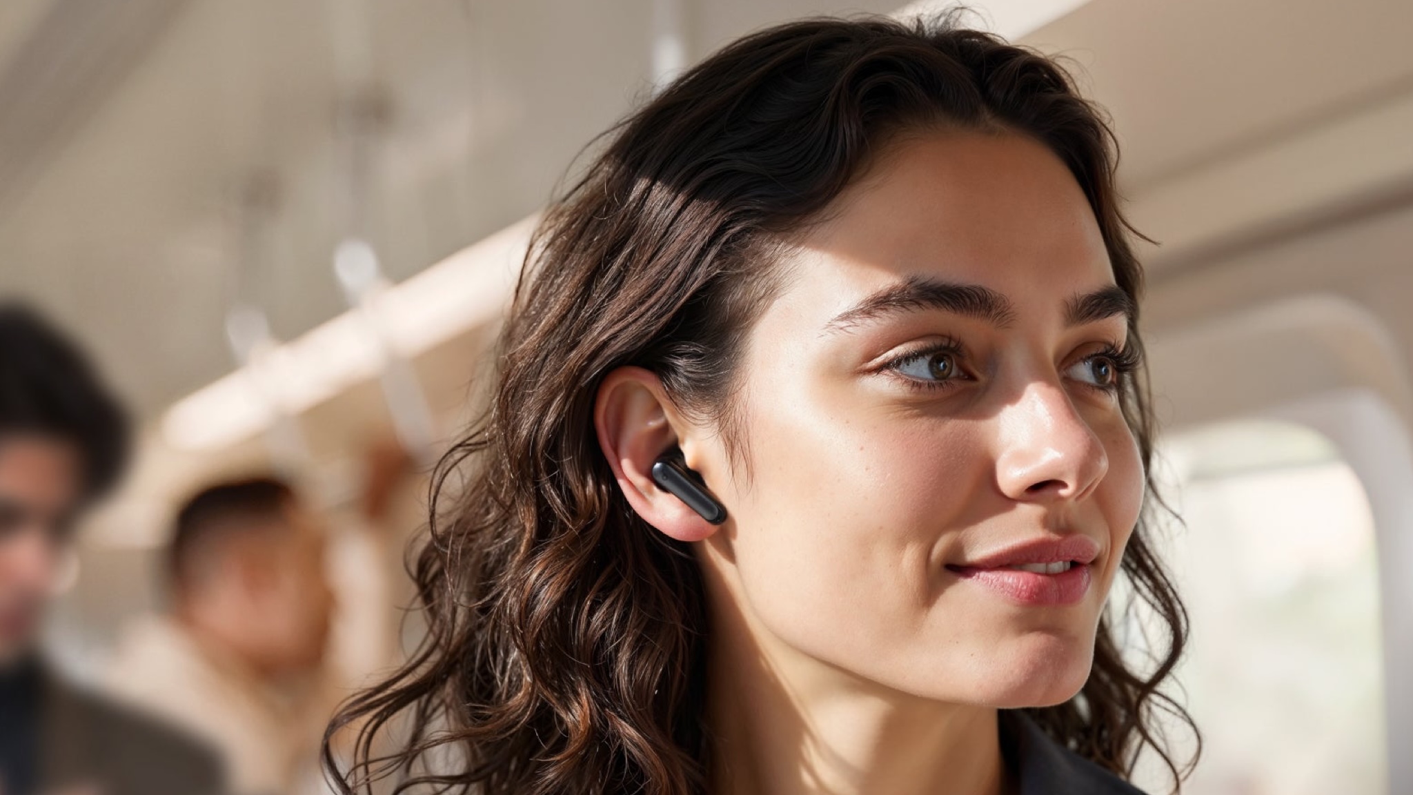 A young woman with brown hair wearing a blue/gray jacket over a white top is on a train or tube with a soundcore P41i earbud in her ear.