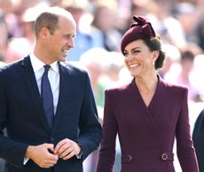 Kate Middleton wearing a purple coat and hat smiling at Prince William wearing a blue suit