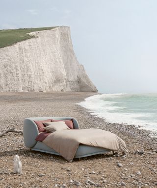 pale blue upholstered bed with peach coloured bedlinen on a pebble beach with cliffs