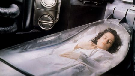 Sigourney Weaver (a white woman with curly brown hair) lies in a bed covered by a clear cover; she appears asleep and is wearing a white night gown