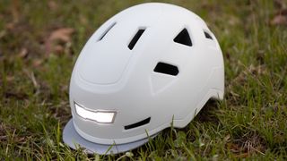 A white Xnito e-bike helmet sits on grass, with the front light illuminated