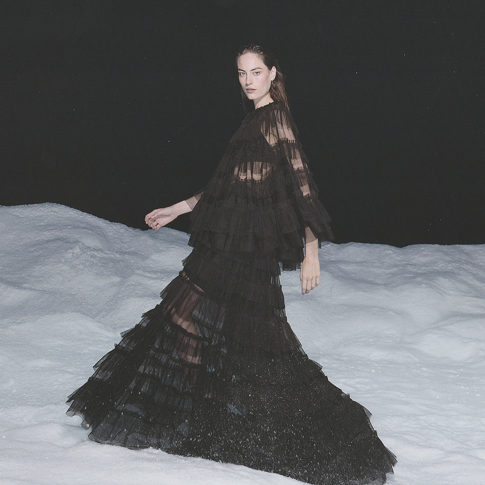 Model walking through snow wearing a black sheer tiered maxi dress sold at AllSaints