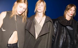 Three male models wearing looks from the Rick Owens collection. One model is wearing dark bottoms and a grey piece that exposes the arm and part of the chest. Another model is wearing a grey coat with a grey piece underneath that exposes parts of the chest and features a chain and straps. And the third model is wearing a black top and black oversized coat. They are standing in front of a blue wall