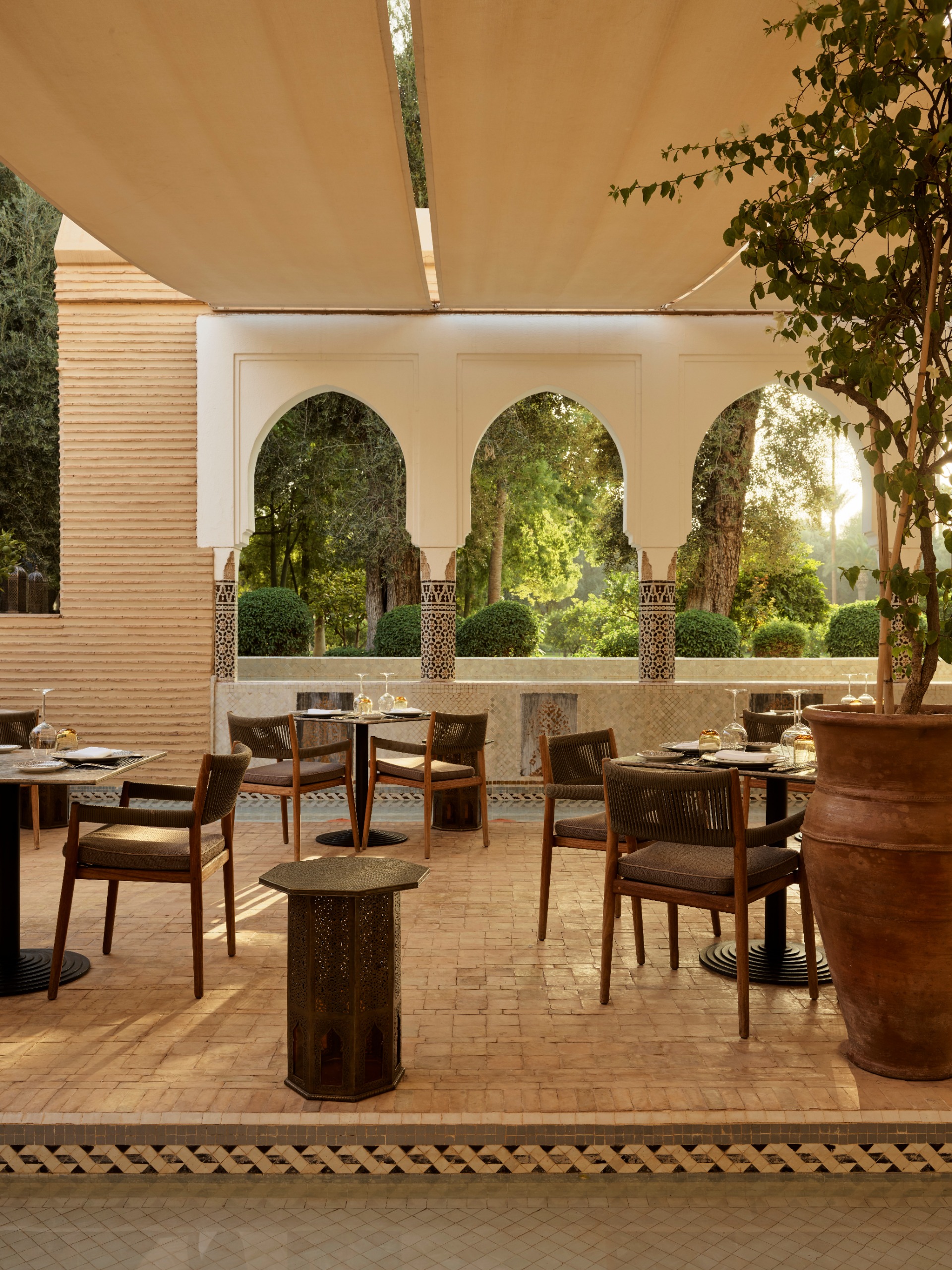 An outdoor restaurant terrace with argilla tiled floors, wooden furniture, large ceramic vases, a lush plant, and arched walls overlooking greenery.