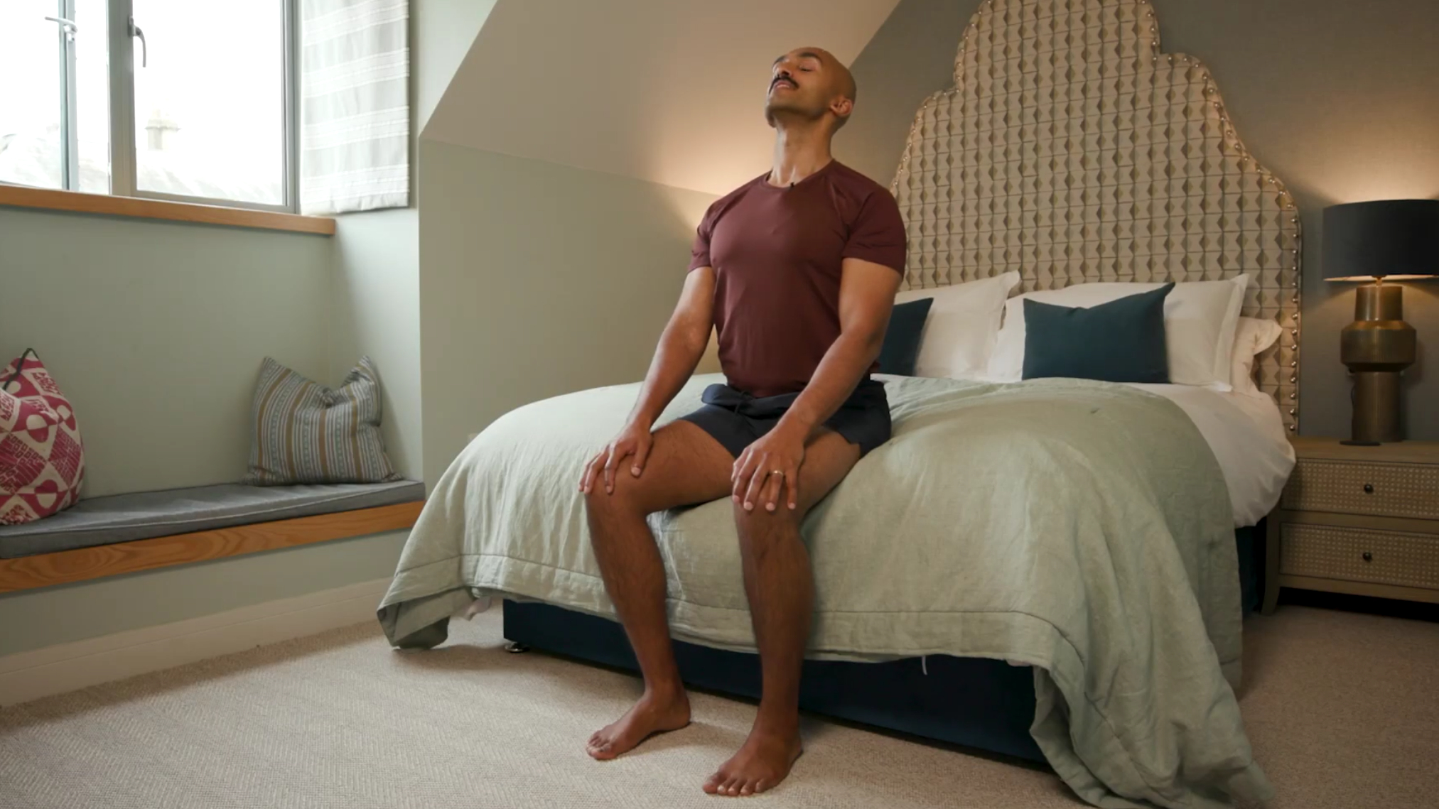 Man sits on edge of bed with his hands on his knees and his head tilted back with his eyes closed