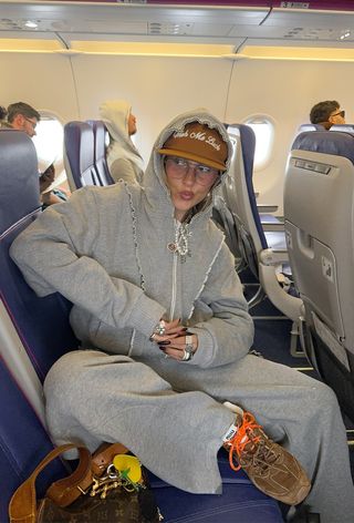 A winter travel outfit is shown in a photo of a woman sitting inside an airplane wearing a tan hat under a gray hoodie styled with matching sweatpants, tan suede sneakers, layered silver rings, and layered necklaces