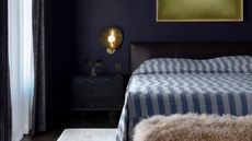 navy bedroom with navy grasscloth wallpaper, green artwork, a brass sconce light, a dark timber side table, a navy chambray bed cover with a fluffy ottoman at the end of the bed