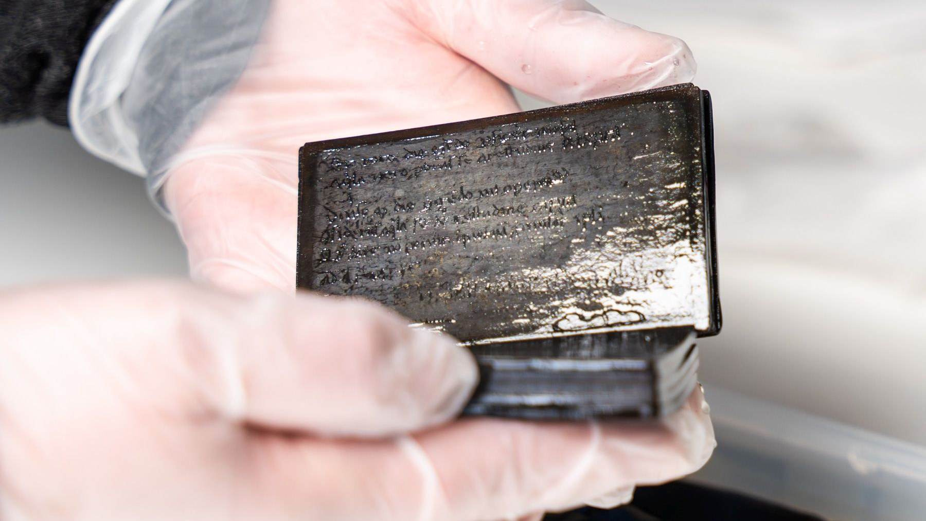 a light-skinned person with clear-gloved hands holds a small wooden tablet with wax inside