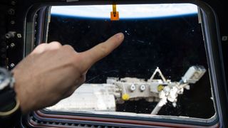 hand with watch points at a divot in a window in space. behind is hardware from the international space station. above is the horizon of earth