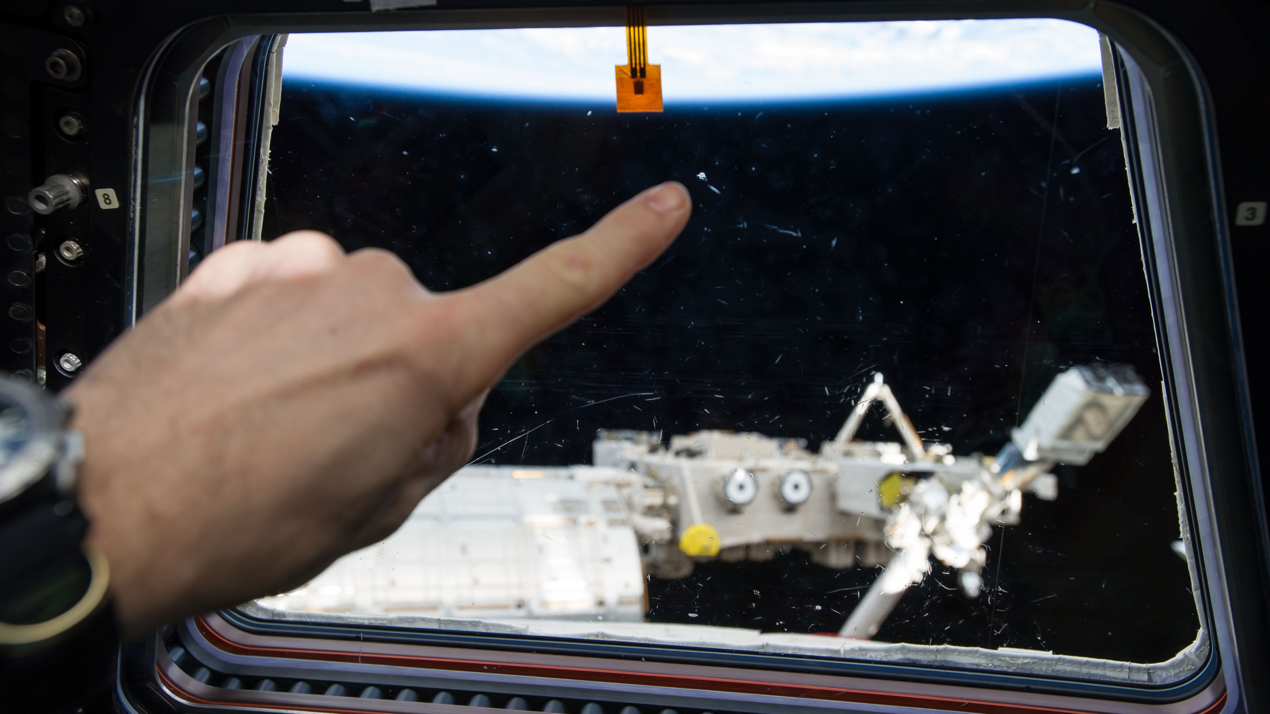 An astronaut on Expedition 40 points to the mark a meteoroid left behind on the International Space Station wraparound cupola window in 2014.