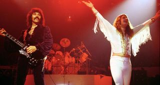 Black Sabbath bathed in red stage lights as they play Madison Square Garden in the late '70s. Tony Iommi is in black. Ozzy Osbourne in white, with white tasselled sleeves and arms aloft.