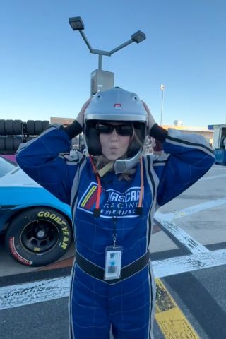 Sydney Sweeney wearing blue NASCAR overalls, holding her helmet and puckering for the camera in front of the famous Concord racetrack.